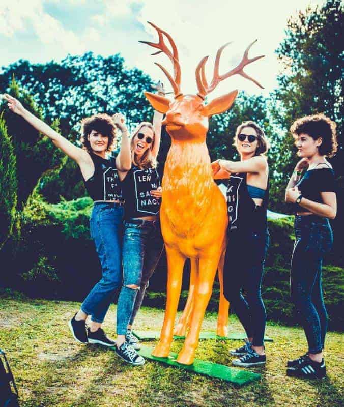 Jeleń Jägermeister 2 Cztery kobiety pozują przy pomarańczowej figurze jelenia na tle zieleni.