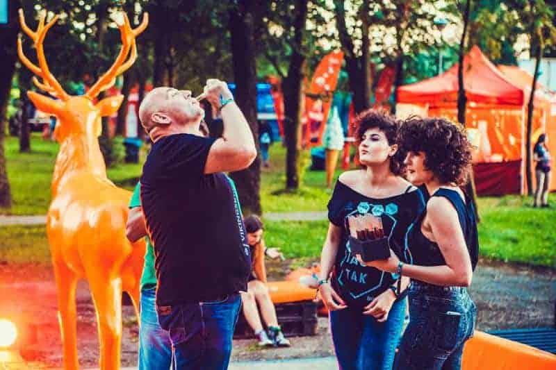 Jeleń Jägermeister 4 Ludzie na festiwalu na świeżym powietrzu, z dużą pomarańczową figurą jelenia w tle, w otoczeniu drzew i stoisk.