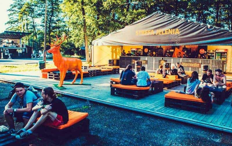 Jeleń Jägermeister 1 Ludzie siedzący na pomarańczowych poduszkach na drewnianej platformie przed namiotem z napisem
