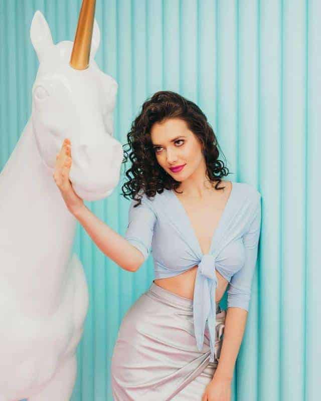 Kobieta w błękitnej bluzce i srebrnej spódnicy, stojąca obok białej rzeźby jednorożca na tle turkusowej ściany.