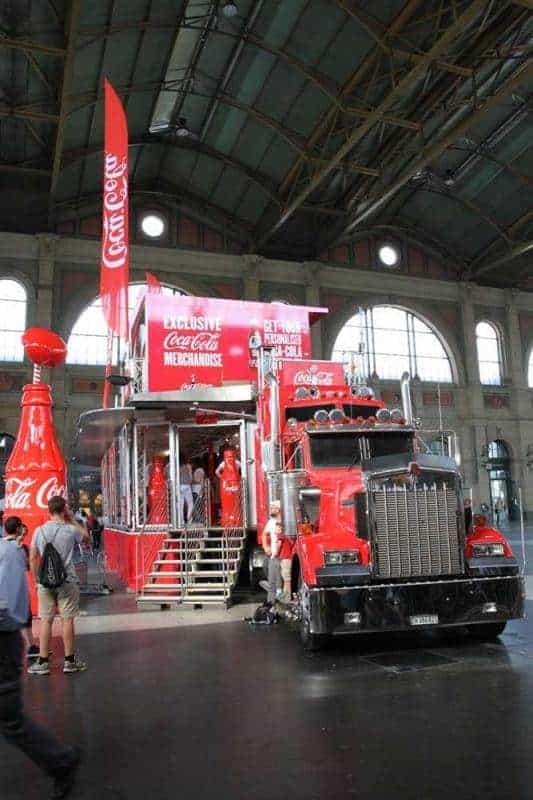 100 lecie szklanej butelki Coca Cola 3 Czerwony ciężarówka Coca-Cola z naczepą przekształconą w stoisko sprzedażowe z produktami Coca-Cola, stojąca w dużej hali.
