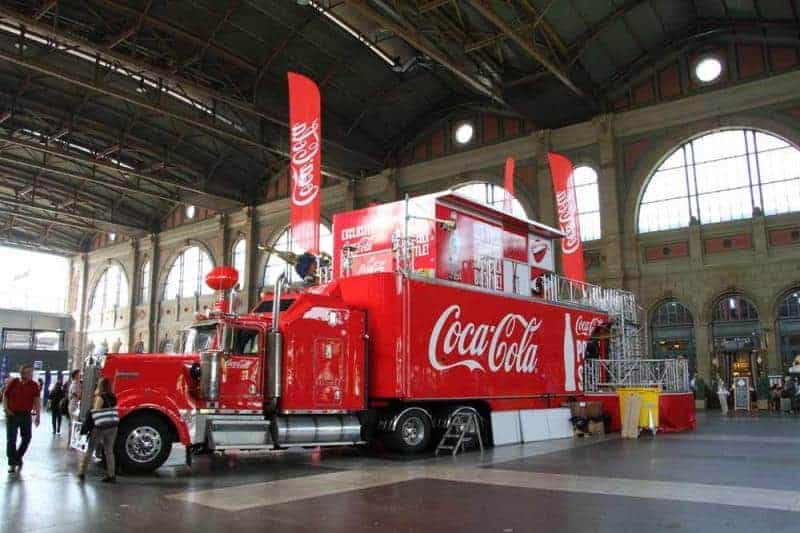 100 lecie szklanej butelki Coca Cola 4 Czerwony ciężarówka Coca-Cola w hali dworca z dużymi flagami Coca-Cola i dużymi oknami w tle.