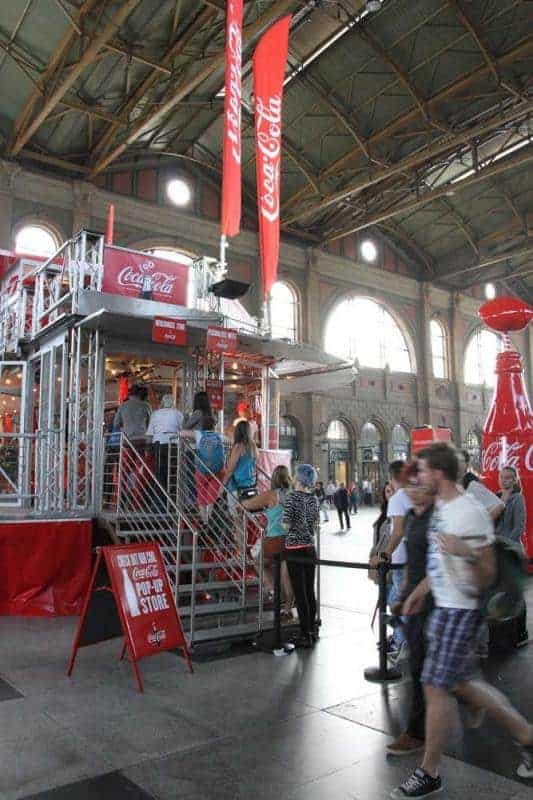 100 lecie szklanej butelki Coca Cola 2 Stoisko Coca-Coli z ludźmi w hali dworca kolejowego.