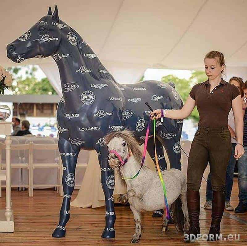 Kobieta trzyma miniaturowego konia na kolorowej smyczy obok dużej statuy konia pokrytej logo Longines.