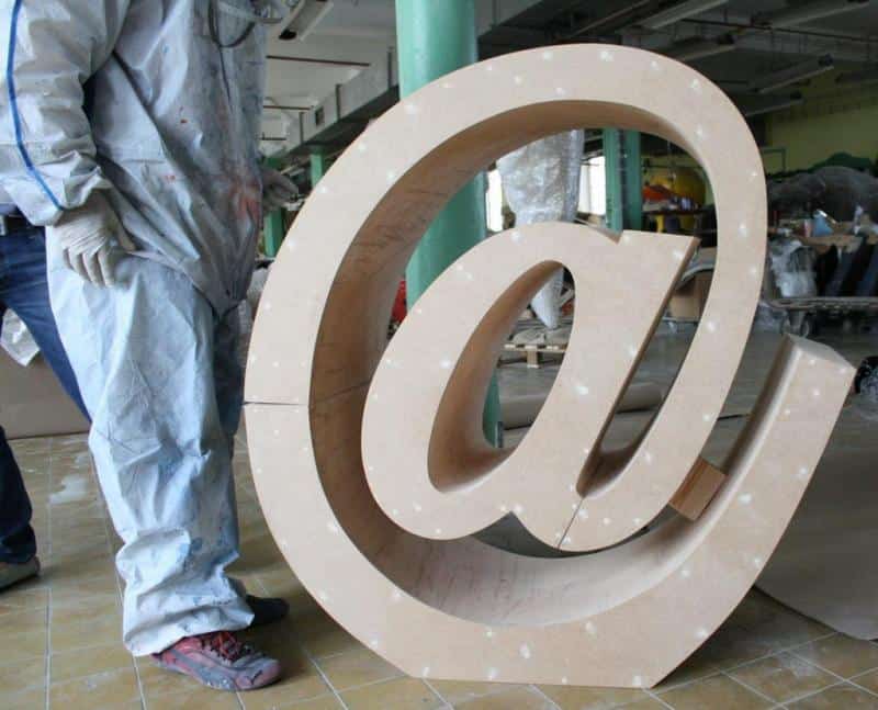 Znak pisarski e-mail 2 Duża rzeźba symbolu małpy (@) w warsztacie, obok której stoi osoba w kombinezonie ochronnym.