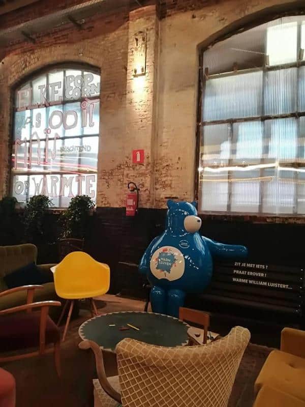 oryg_064490685_1129949550544944_1465385417682452480_o Pomieszczenie z ceglaną ścianą, dużymi oknami, kolorowymi krzesłami oraz niebieską figurą misia siedzącą na ławce.