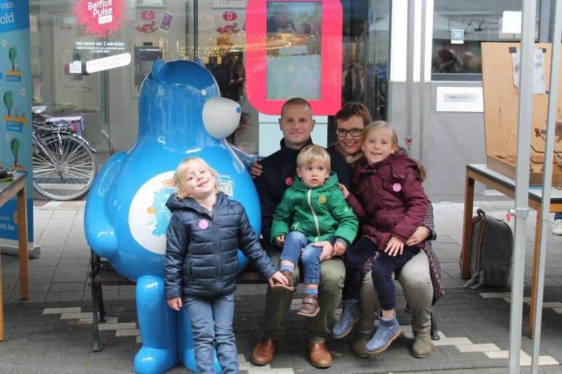 Miś na ławce 2 Rodzina z trójką dzieci pozuje do zdjęcia obok dużej niebieskiej maskotki w przestrzeni publicznej.