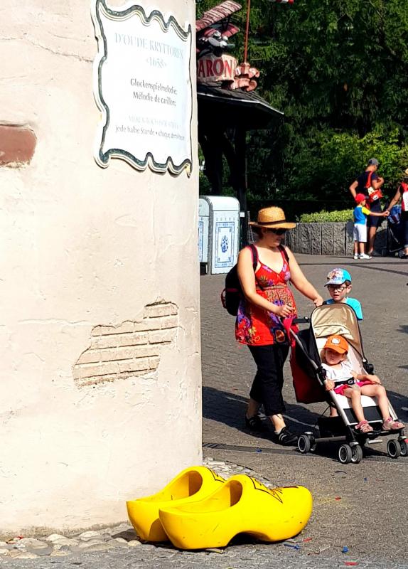 Wielkie chodaki 4 Kobieta z wózkiem dziecięcym przechodzi obok dużych żółtych drewniaków przy ścianie z francuskim napisem.