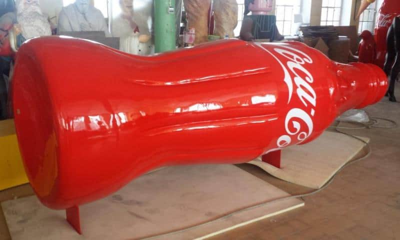 Duża rzeźba w kształcie butelki Coca-Coli w czerwonym kolorze, znajdująca się w warsztacie.