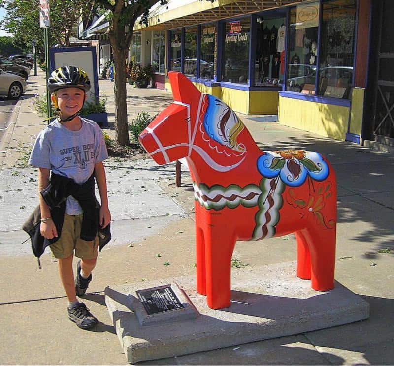 Artystycznie malowane konie z Dalarna 2 Chłopiec w kasku na rower stojący obok kolorowej rzeźby konia na chodniku wzdłuż ulicy z witrynami sklepów.