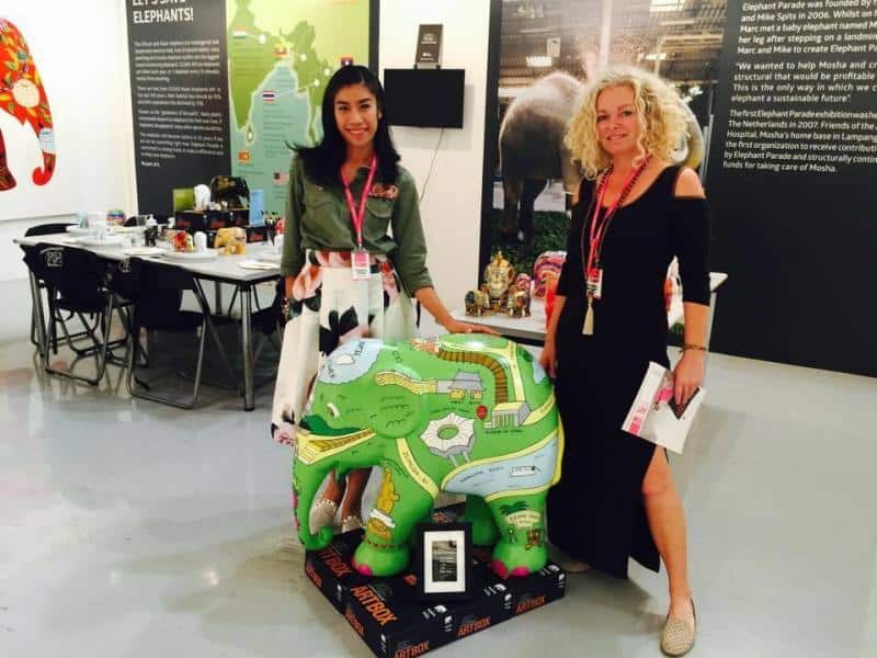 Dwie kobiety stojące obok kolorowej figurki słonia, trzymające broszury na tle wystawy.