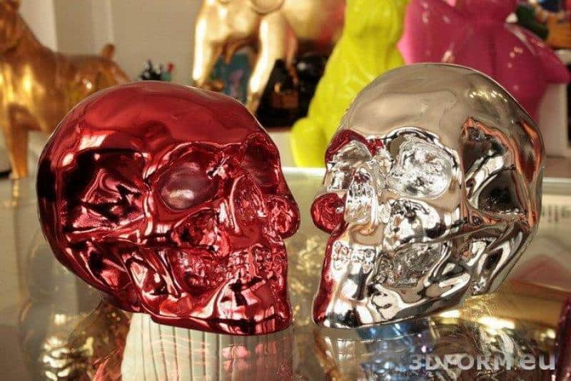 Dwie metaliczne czaszki, jedna czerwona, druga srebrna, ustawione na szklanym stole.