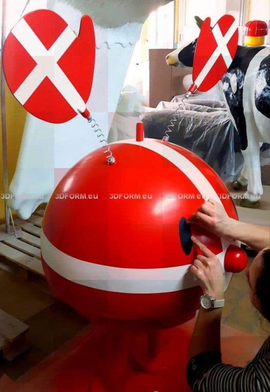 Figura reklamowa Hoptimist 1 Czerwona, okrągła figura z uszami na sprężynach i białymi paskami, z rękami przymocowującymi oko. W tle plastikowa krowa.