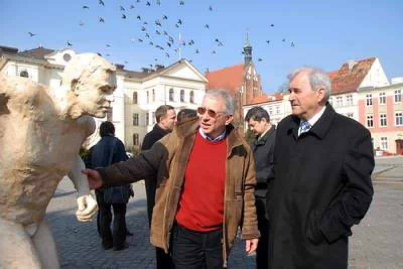 Promocja mistrzostw świata IAAF 1 Dwóch mężczyzn rozmawiających obok rzeźby na rynku miejskim, w tle kamienice i wieża zegarowa.