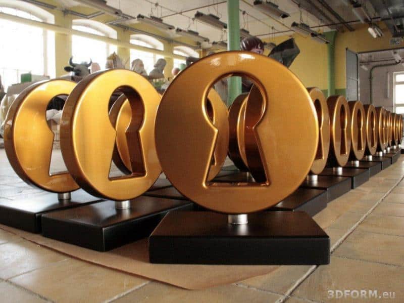 Złote statuetki w kształcie zamka na czarnych podstawach ustawione w rzędzie w przestrzeni przemysłowej.