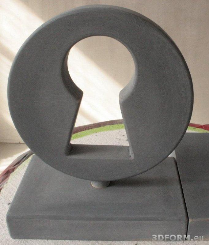 Szara rzeźba w kształcie zamka do klucza, umieszczona na prostokątnej podstawie.