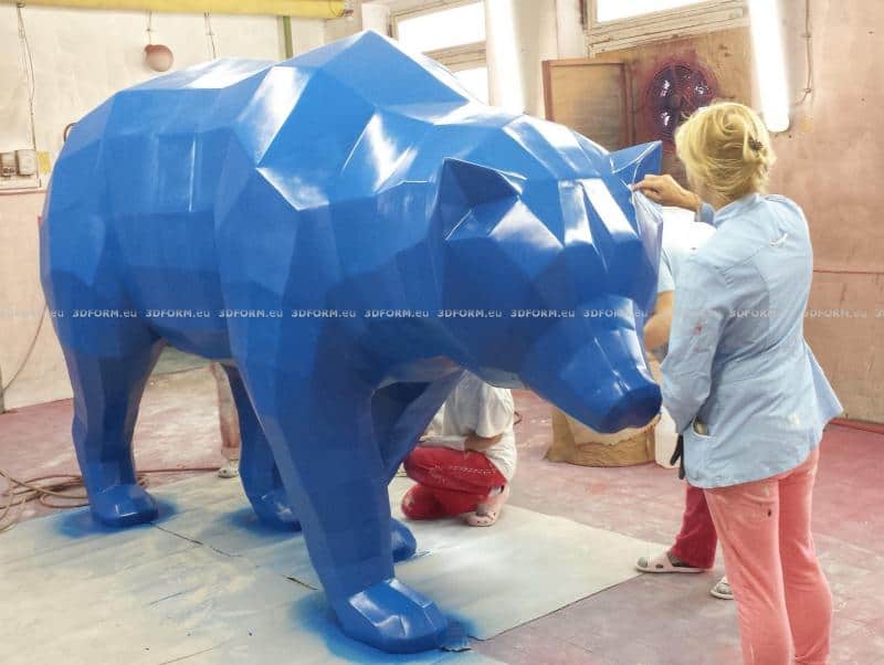 Geometryczny niedźwiedź typu low-poly 1 Kobieta maluje dużą, niebieską, geometryczną rzeźbę niedźwiedzia w pracowni artystycznej.