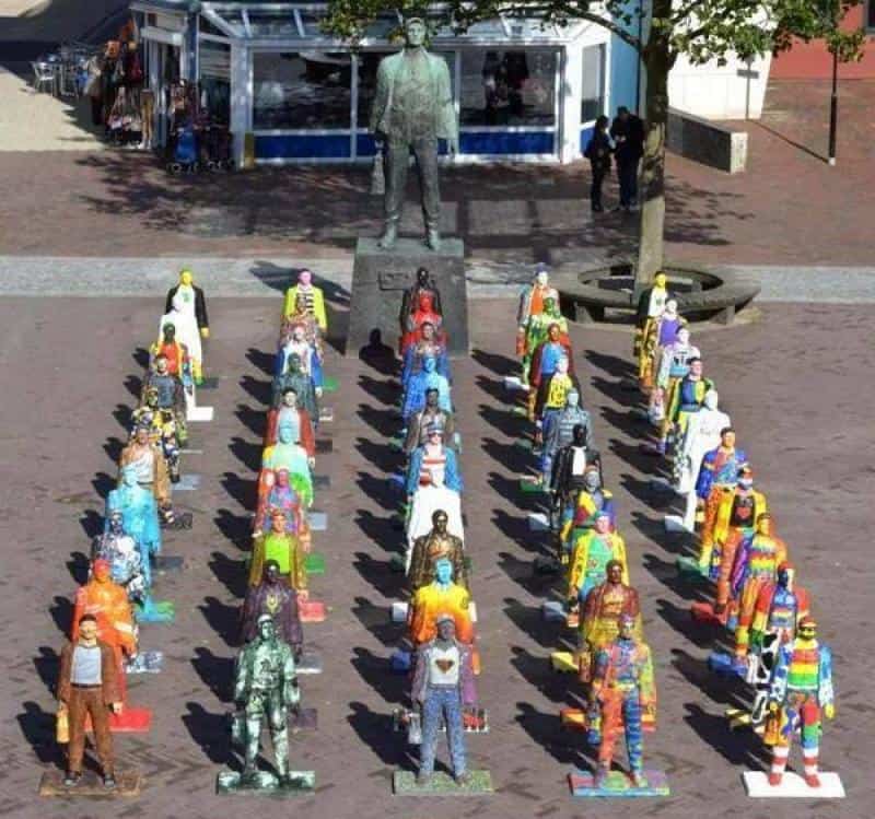 Rzeźby górników 1 Kolorowe figury ludzi ustawione w szeregu na placu przed dużym pomnikiem mężczyzny.