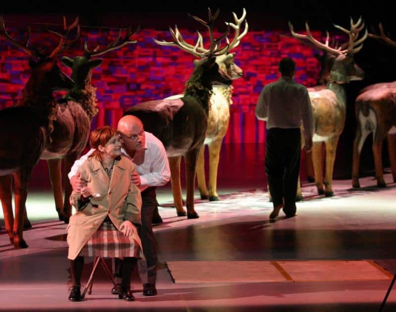 oryg_064490685_1129949550544944_1465385417682452480_o Scena teatralna z aktorami i plastikowymi jeleniami na tle czerwonej dekoracji.