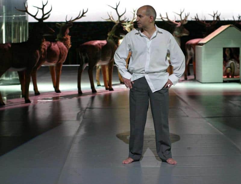 Elementy scenografii dla Teatru Dramatycznego Miasta Stołecznego Warszawy 1 Mężczyzna w białej koszuli i szarych spodniach stoi boso na scenie, w tle stoją wypchane jelenie.