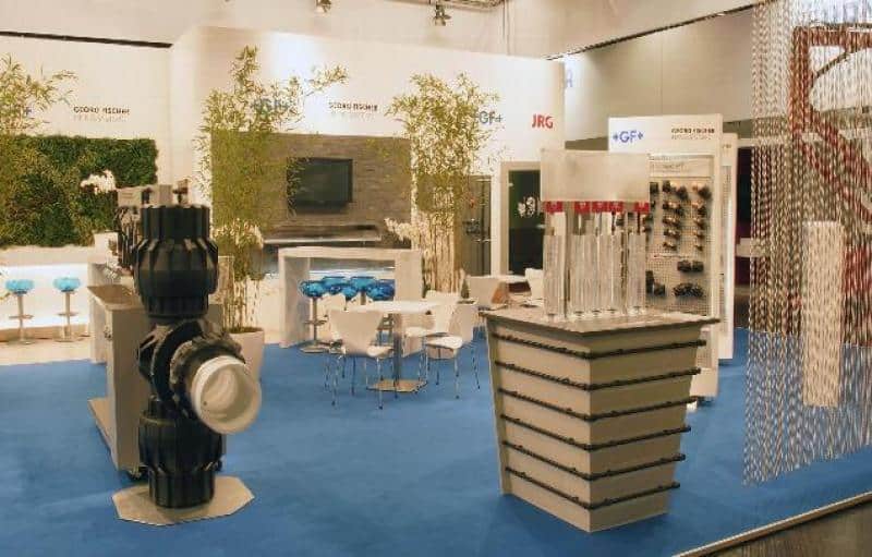 oryg_064490685_1129949550544944_1465385417682452480_o Stoisko wystawowe z armaturą przemysłową, białymi stolikami i krzesłami na niebieskim dywanie.