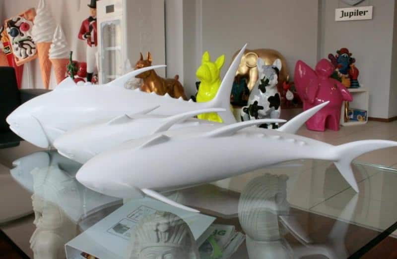 Ławica tuńczyków 3 Białe figurki rekinów na szklanym stole, w tle kolorowe figurki zwierząt i inne dekoracje.