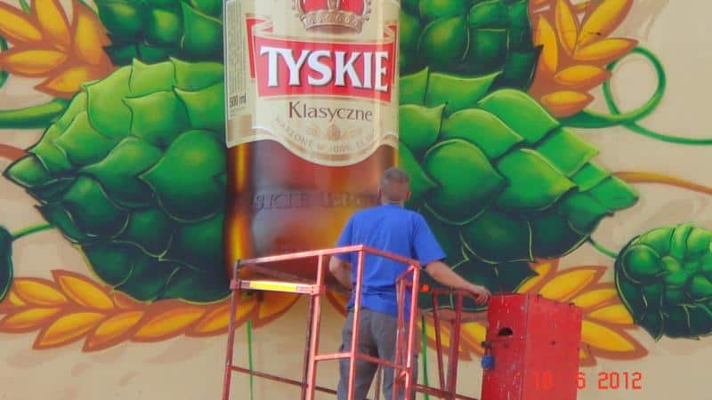 Trójwymiarowy mural TYSKIE 4 Mężczyzna na podnośniku malujący mural z butelką piwa Tyskie Klasyczne i chmielami.