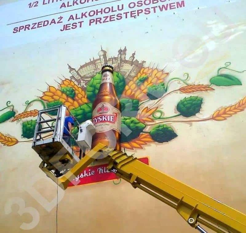 Trójwymiarowy mural TYSKIE 2 Osoba na podnośniku malująca reklamę piwa Tyskie na ścianie budynku.