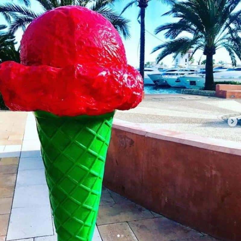 Makieta loda - Zielona Budka 4 Wielka plastikowa rzeźba przedstawiająca loda rożka z czerwoną gałką na zielonym wafelku, umieszczona na zewnątrz w otoczeniu palm i jachtów.