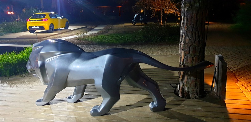 Figurka lwa w kolorze srebrnym na drewnianej platformie, w tle żółty samochód na oświetlonym podjeździe.