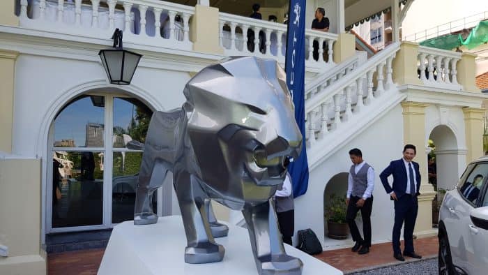 Metalowy posąg lwa na tle białego budynku z kolumnadą i schodami, przy którym stoją elegancko ubrani ludzie.