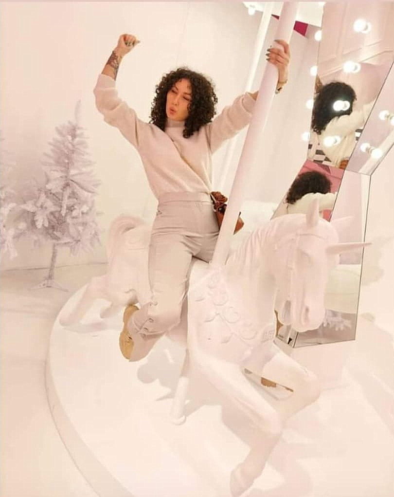 Karuzela wyjątkowych jednorożców 3 Osoba siedząca na karuzeli z białym jednorożcem w jasnym pomieszczeniu, obok znajdują się lustra i ozdobione białe choinki.