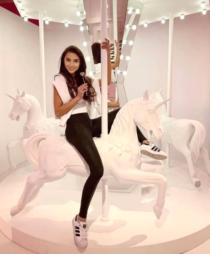 Karuzela wyjątkowych jednorożców 1 Kobieta siedząca na białym jednorożcu na karuzeli, trzymająca lizaka, otoczona jasnymi światłami.