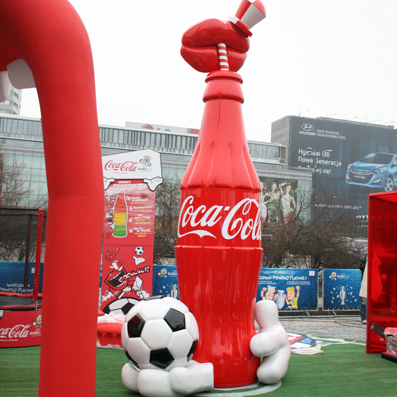 Nadmuchiwany model butelki Coca-Coli obok dużej nadmuchiwanej piłki nożnej na zewnątrz budynku.