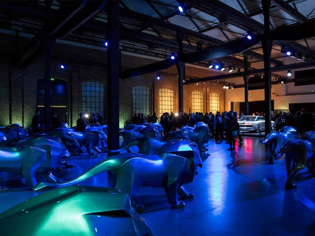 Pomieszczenie wypełnione niebieskimi rzeźbami lwów, z ludźmi i samochodem w tle, oświetlone niebieskim i fioletowym światłem.