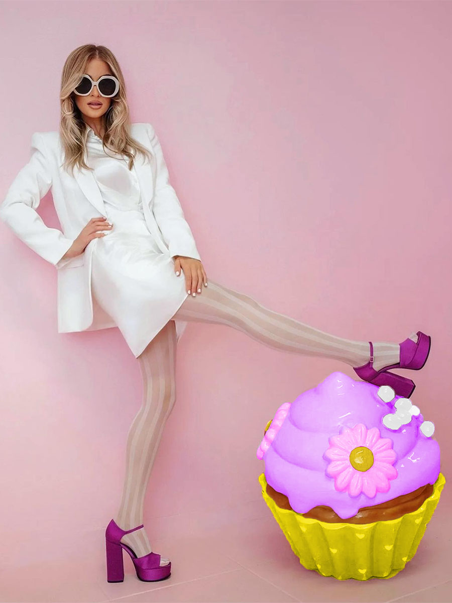 Kolorowa Babeczka dla Cukierni Sweet Home w Lagos, Portugalia 4 Kobieta w białym stroju, okularach przeciwsłonecznych i fioletowych butach na wysokim obcasie na tle różowej ściany, z fioletowo-żółtą dekoracyjną babeczką.