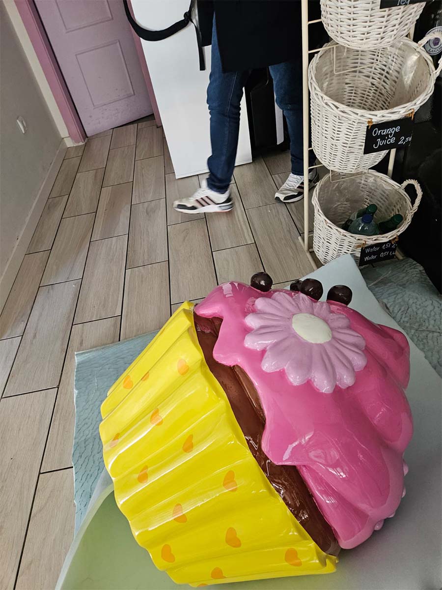 Kolorowa Babeczka dla Cukierni Sweet Home w Lagos, Portugalia 3 Rzeźba wielkiego babeczki w pastelowych kolorach stojąca na podłodze w kawiarni obok osoby w dżinsach i białych butach.