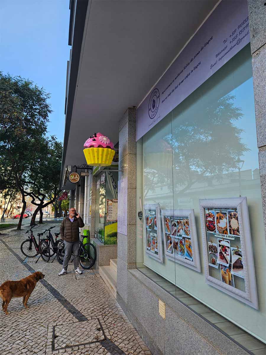 oryg_064490685_1129949550544944_1465385417682452480_o Mężczyzna na chodniku przed sklepem z różowym szyldem w kształcie muffinki, z psem i rowerami obok.