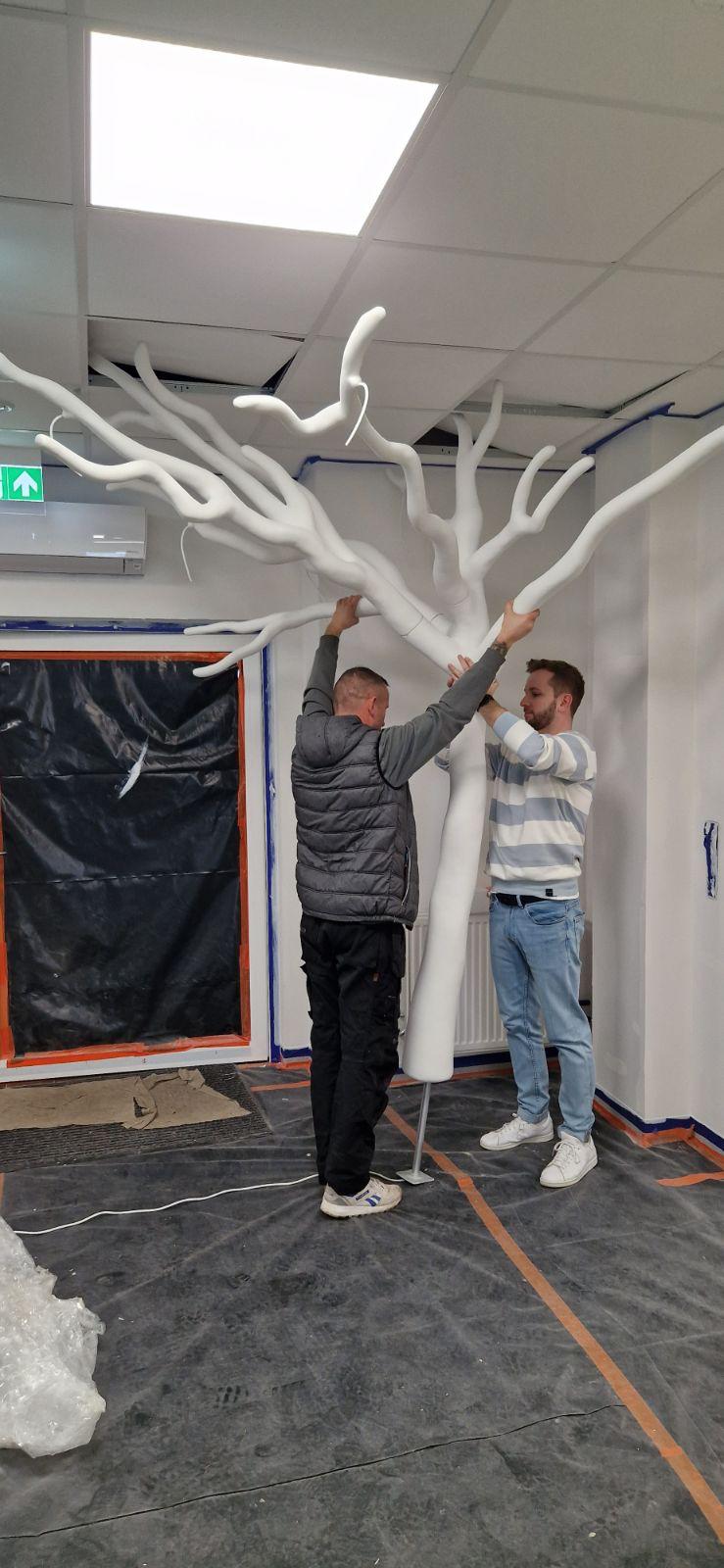 Dwóch mężczyzn montujących dużą, białą rzeźbę drzewa w pomieszczeniu.