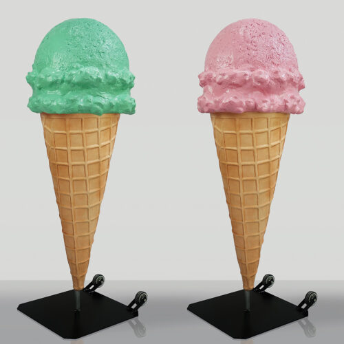 Dwa duże, plastikowe lody w wafelkach na stojakach. Po lewej lód pistacjowy, po prawej truskawkowy.