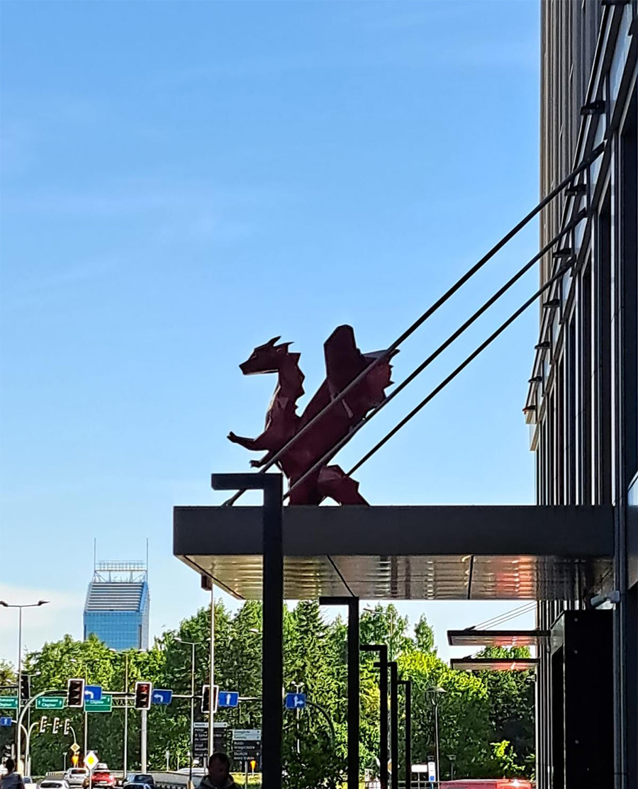 Czerwony smok na daszku budynku, w tle miejski krajobraz z drzewami i budynkami.