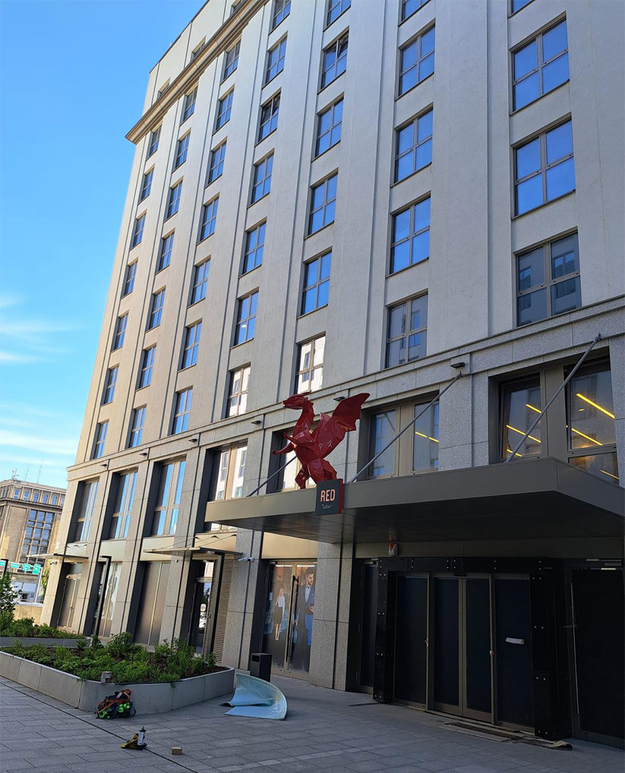 Wysoki budynek z szarymi ścianami i licznymi oknami, przed wejściem czerwony posąg smoka z napisem 