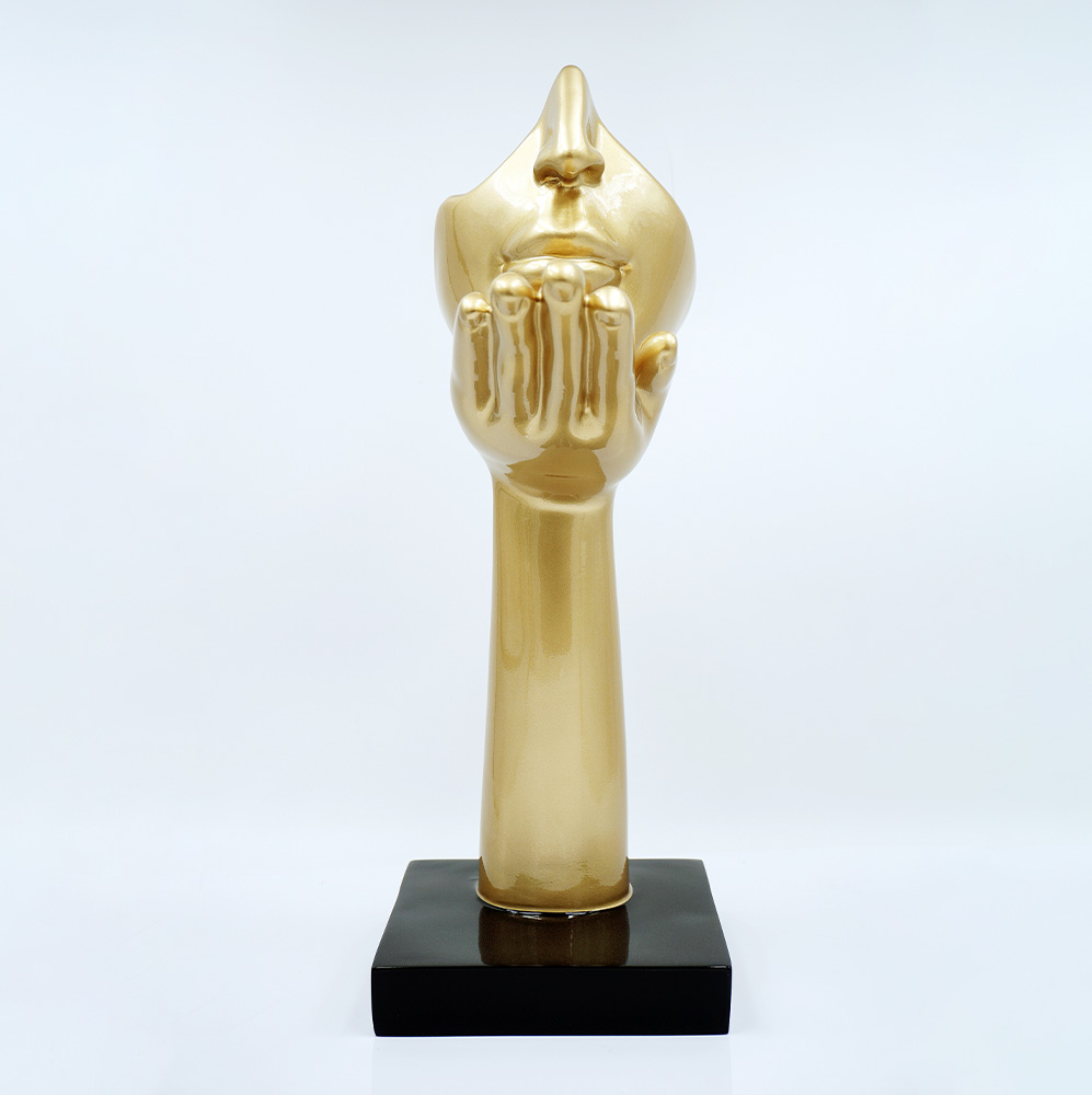 Mała Maska Na Podstawie - Motyw I 2 Złota rzeźba przedstawiająca dłoń wzniesioną do ust twarzy o zamkniętych oczach, na czarnej podstawie, na białym tle.