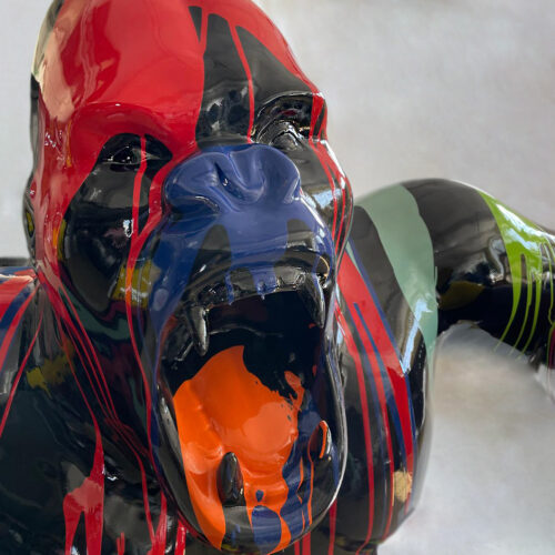 Rzeźba goryla z otwartą paszczą, pomalowana kolorowymi, błyszczącymi farbami w czerwonym, czarnym, niebieskim, zielonym i pomarańczowym kolorze, na tle jasnej ściany.