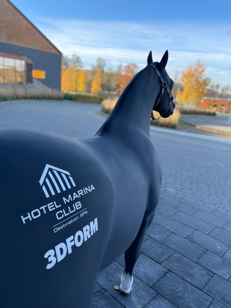 Figura czarnego konia z białym napisem HOTEL MARINA CLUB Destination SPA 3DFORM na grzbiecie, stojąca na bruku na tle jesiennego krajobrazu z budynkami i drzewami o żółtych liściach