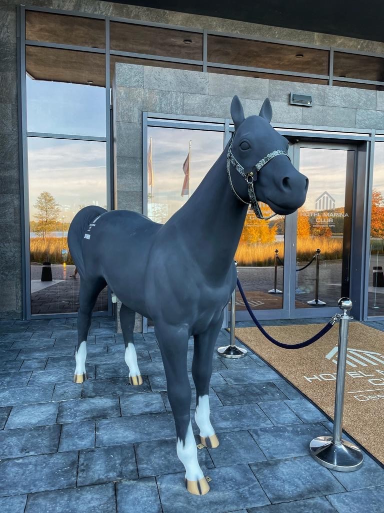 Rzeźba czarnego konia z białymi kopytami stojąca przed budynkiem hotelu z szarymi kamiennymi ścianami i dużymi szklanymi drzwiami.