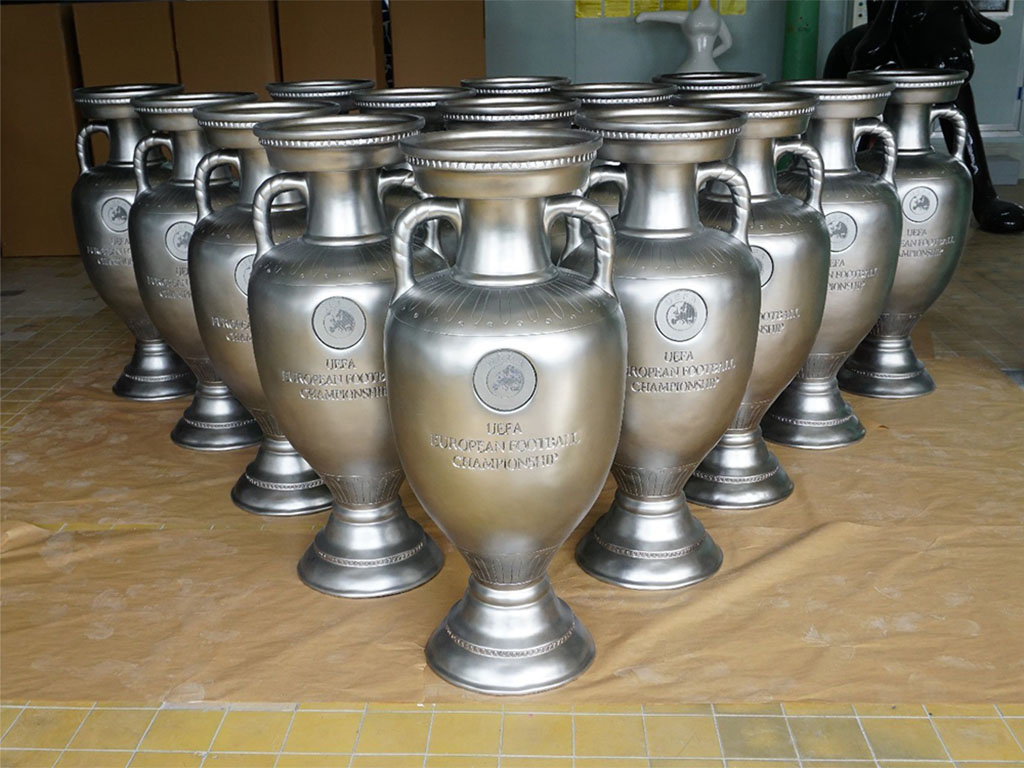Kilka srebrnych trofeów UEFA European Football Championship ustawionych w dwóch rzędach na podłodze wewnątrz pomieszczenia.