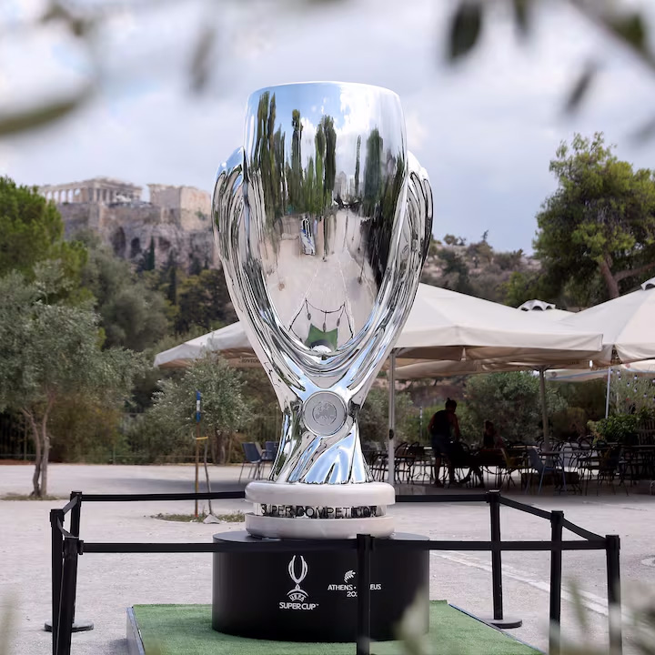 srebrny puchar Superpucharu piłkarskiego na podstawie na zewnątrz, z widokiem na ruiny Akropolu w tle i parasolami kawiarnianymi