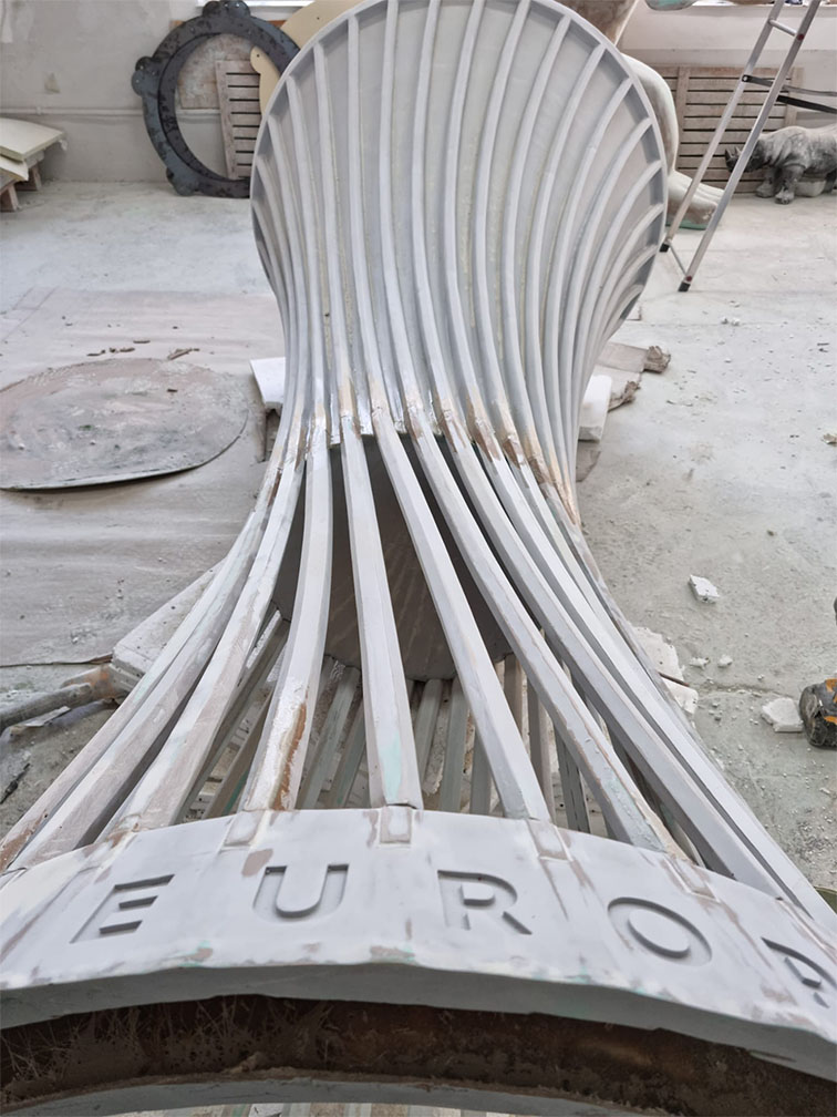 Biały, nowoczesny i zakrzywiony fotel wykonany z cienkich, pionowych listew, leżący na podłodze w surowym pomieszczeniu, z napisem EURO na dole fotela.