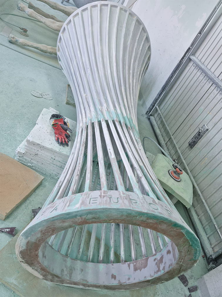 Warsztat z dużą, białą, cylindryczną metalową konstrukcją o smukłym kształcie leżącą na podłodze, z widocznymi rękawicami roboczymi i narzędziami wokół niej.